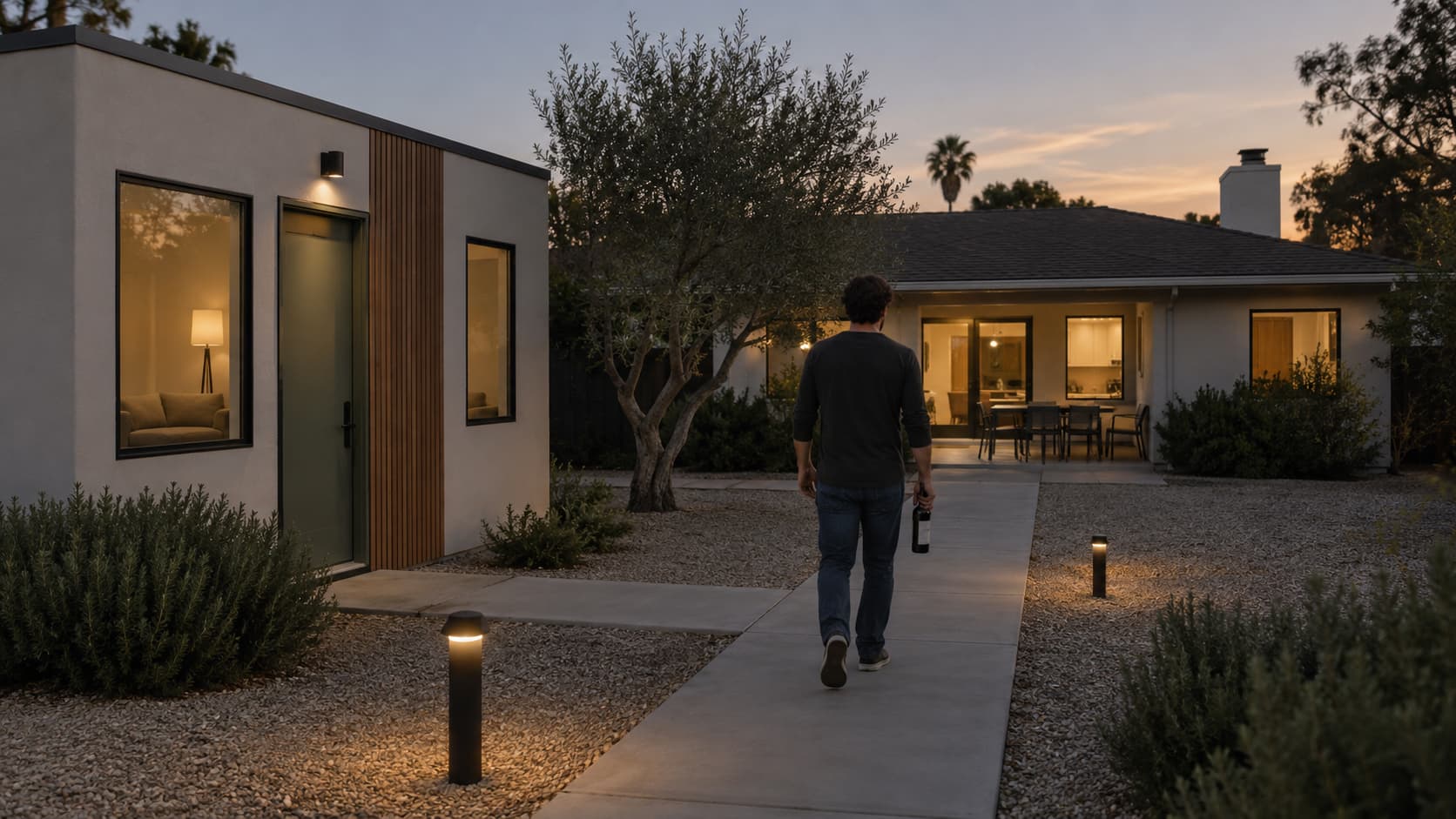 A young man walking along a lit path from his ADU toward the main house carrying a bottle of wine